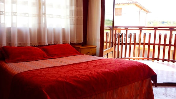 Un dormitorio con una cama grande, ropa de cama roja, una mesita de noche de madera y vistas a un balcón con barandilla.