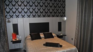Ein modernes Schlafzimmer mit gemustertem Kopfteil, einem Bett mit gestreifter Bettdecke, zwei schwarzen Kissen, einem schwarzen Bademantel, einem Nachttisch mit Lampe und einer kleinen Ablage mit Toilettenartikeln.