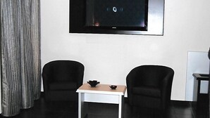 Ein modernes Zimmer mit einem an der Wand befestigten Flachbildfernseher, einem kleinen weißen Tisch mit zwei schwarzen Stühlen und einem Vorhang links.