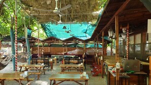 An outdoor dining area with wooden tables and benches, covered by a thatched roof and surrounded by greenery. - Olympos Tatilya Pansiyon (Kumluca)