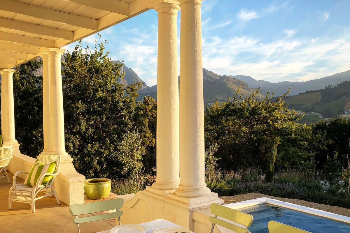 A covered outdoor dining area with white columns, a pool, and a mountainous backdrop.