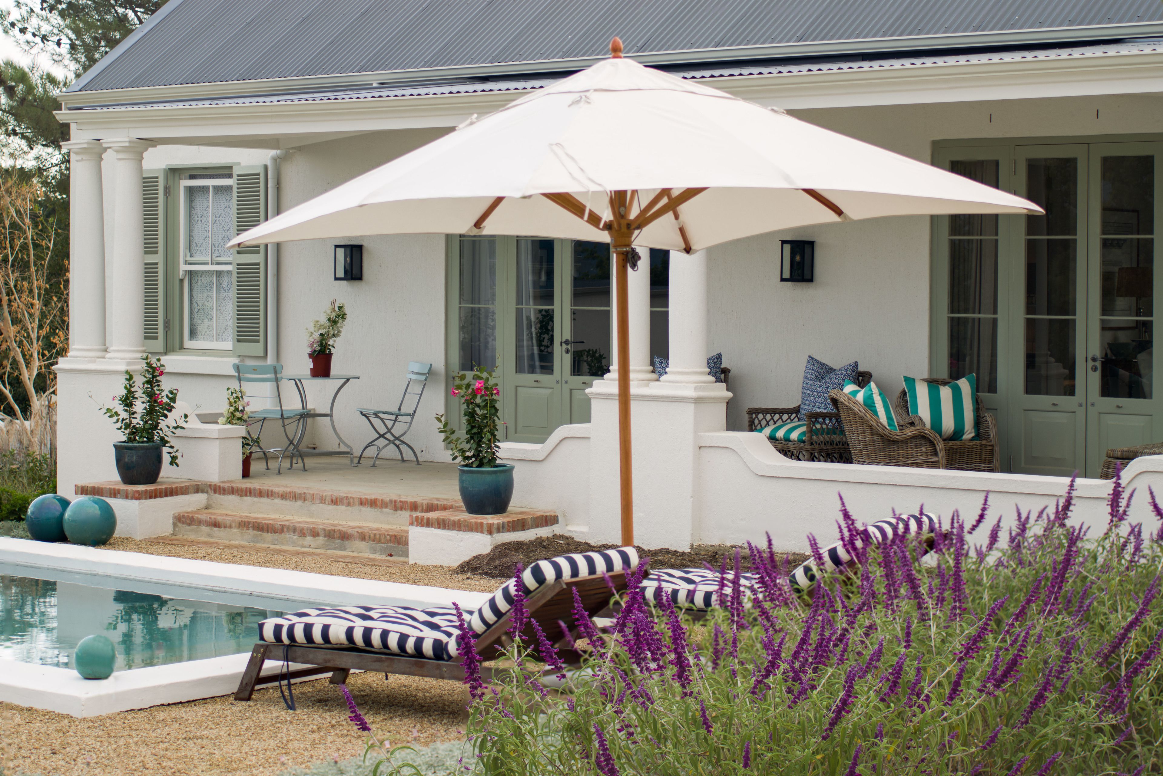 A pool area with lounge chairs and an umbrella, surrounded by potted plants and a house with a porch.