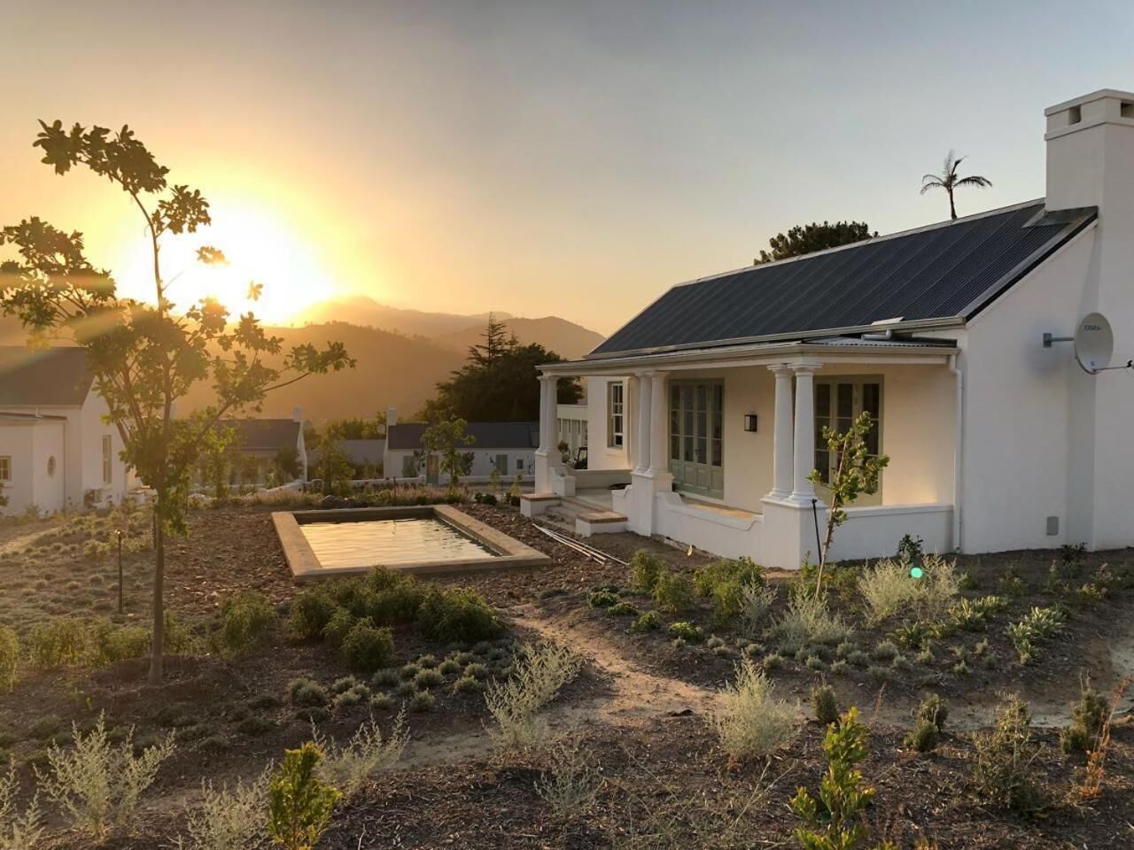 Une maison blanche avec un toit équipé de panneaux solaires, une piscine et un jardin où poussent différentes espèces de plantes.