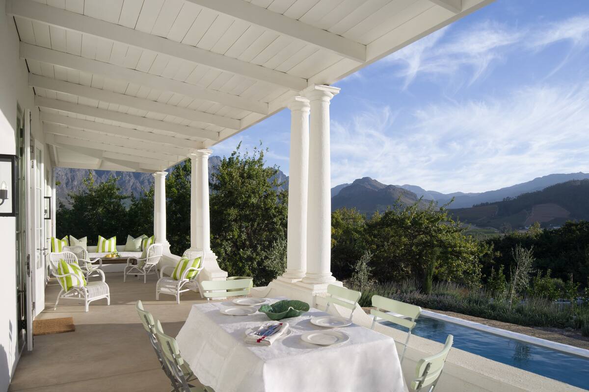 A covered outdoor dining area with white columns, green chairs, and a view of mountains.