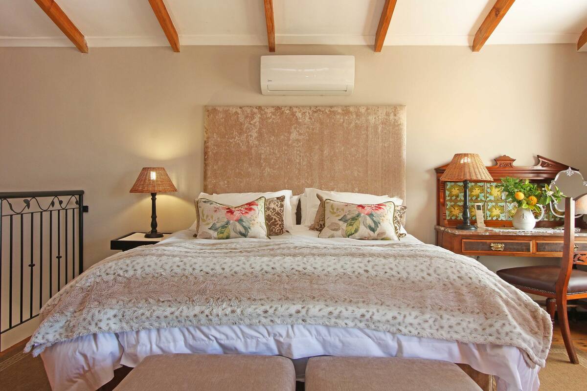A bedroom with a large bed, a nightstand with a lamp, a dresser with a mirror, and an air conditioner on the wall.