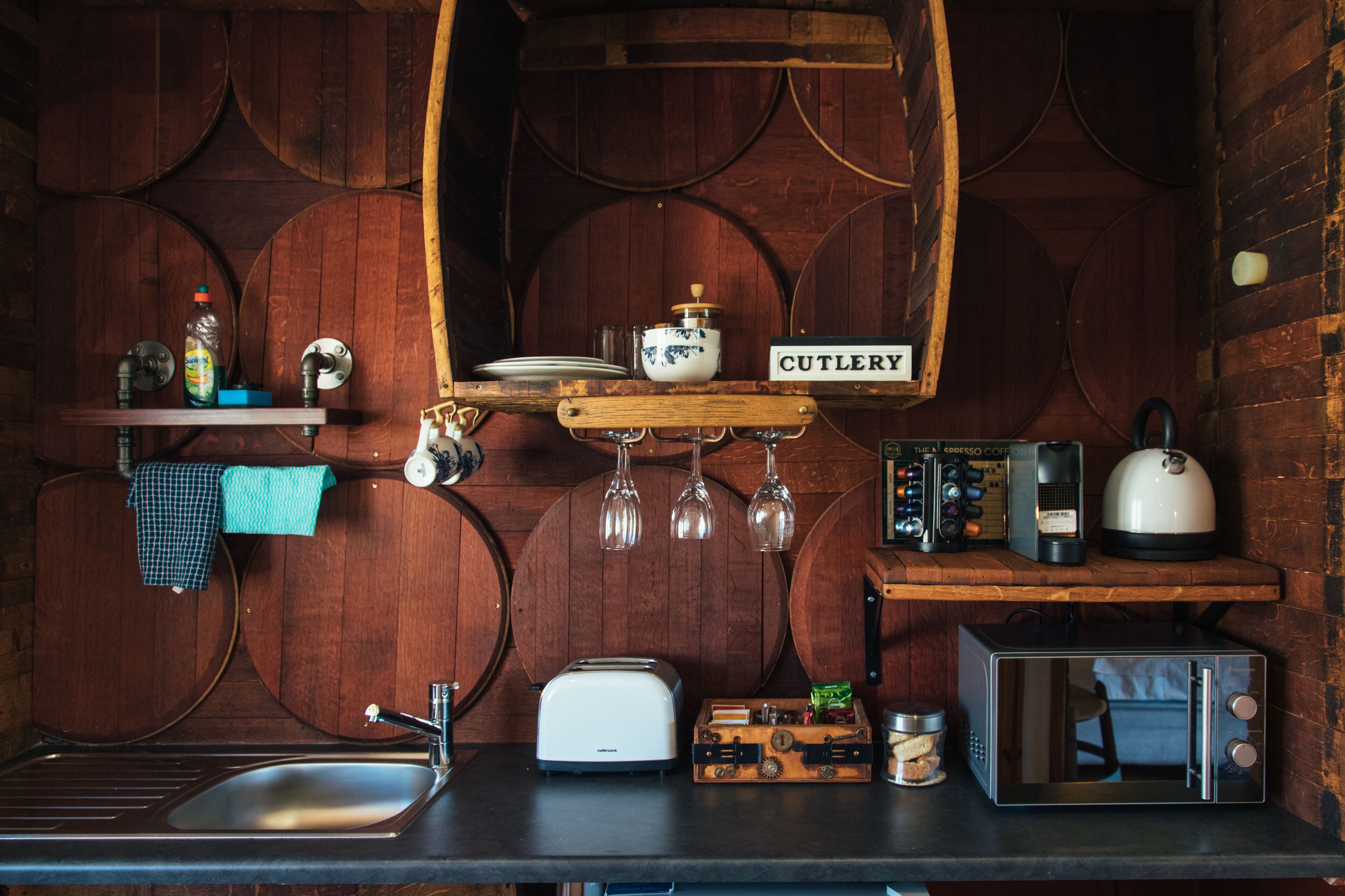 A rustic kitchen with wooden barrels as walls, a cutlery shelf, a toaster, a kettle, and a microwave.