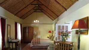 A room with a high wooden ceiling, a bed with a floral bedspread, a bedside table with a lamp, a chair, and a small table with a vase of flowers. - High Hopes (Greyton)