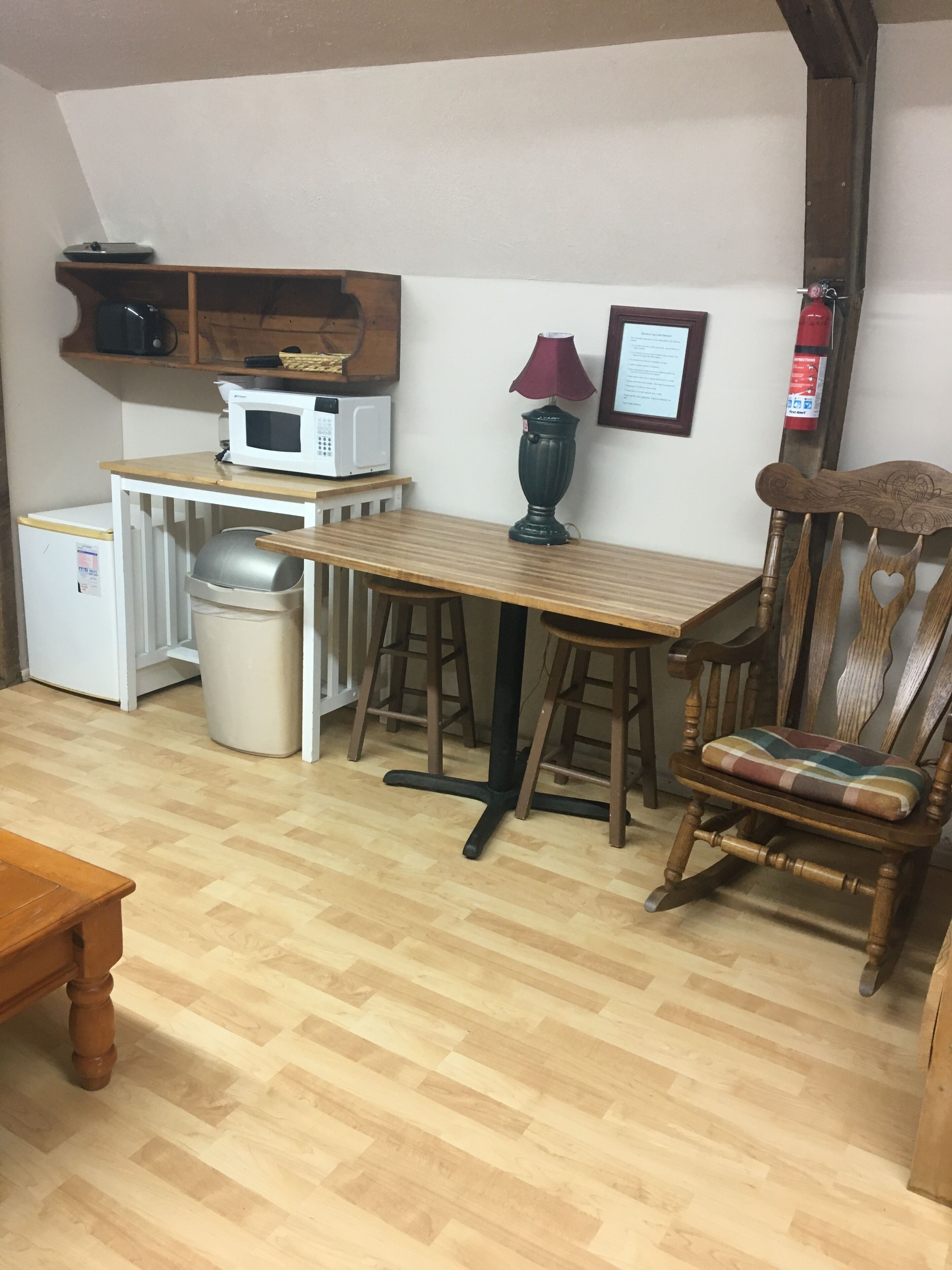 A compact kitchen with a microwave, a small refrigerator, a trash bin, a wooden table, and a chair with a heart-shaped cushion.