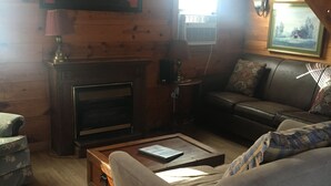 Interior acogedor de una cabaña con chimenea, paredes de madera, un sofá y una ventana.