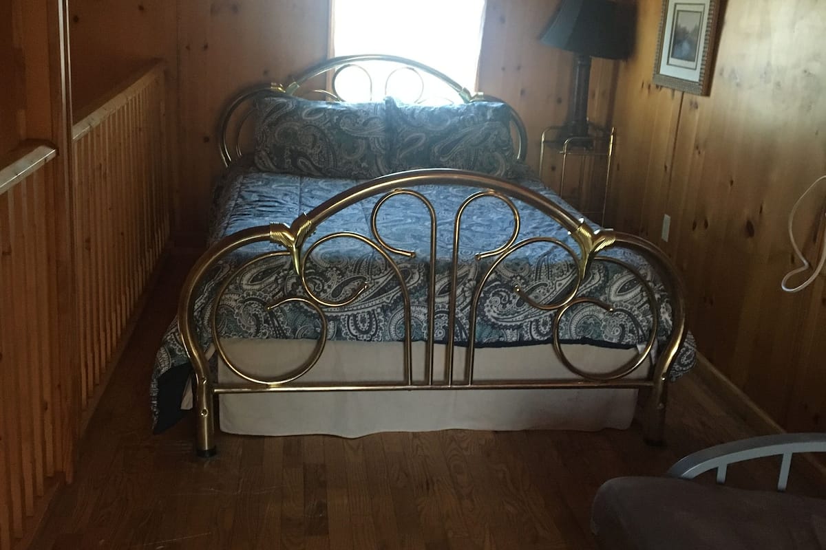 Une chambre dans une cabane en bois, avec un lit en métal, une fenêtre avec des rideaux, une table de chevet avec une lampe et un tableau encadré accroché au mur.