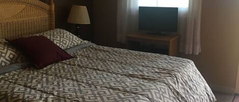 Ein Schlafzimmer mit Bett, Fernseher auf einem Holzschrank, Lampe und einem Fenster mit Vorhängen.