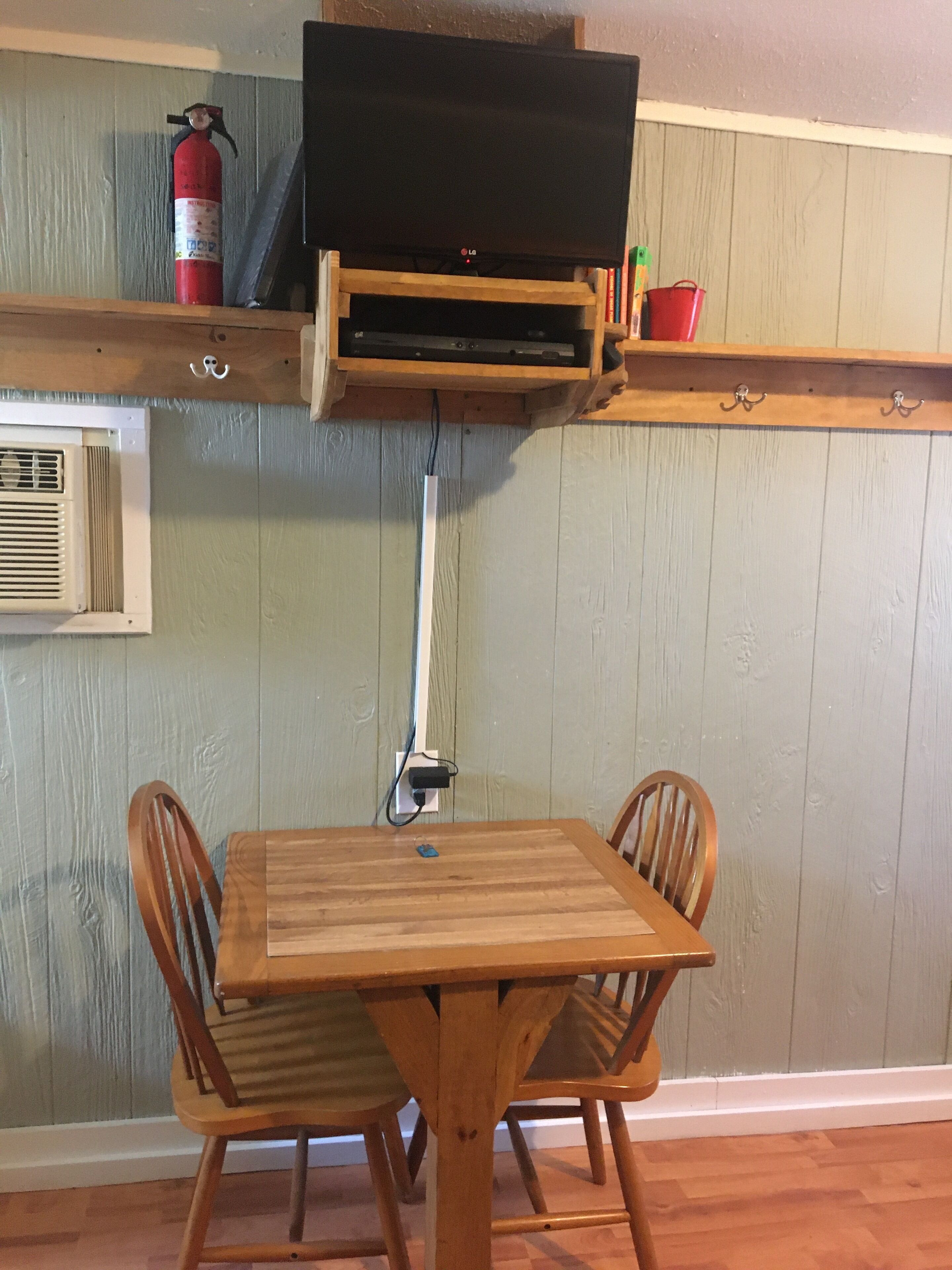 A wooden dining table with two chairs, a television mounted on a wooden shelf above, and a fire extinguisher on the wall.