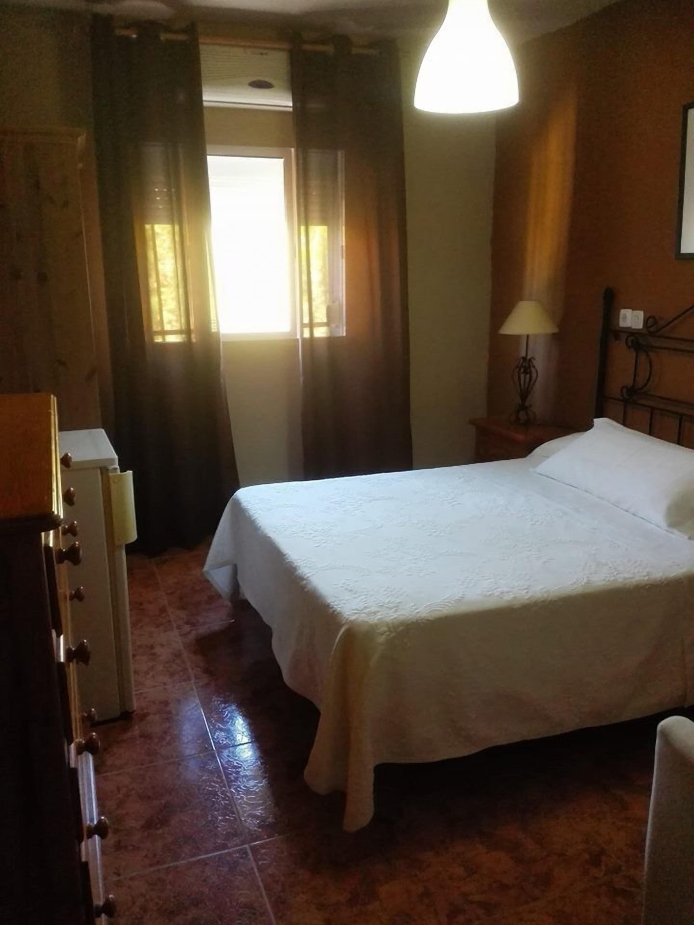 A bedroom with a bed, a nightstand with a lamp, a window with curtains, and a wooden wardrobe.