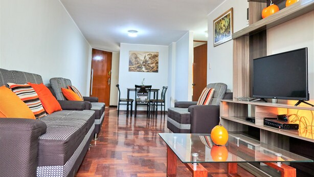 A modern living room with a sofa set, a glass coffee table, a flat-screen TV, and a dining area with chairs and a table. - Eral Apartments Lima (Lima)