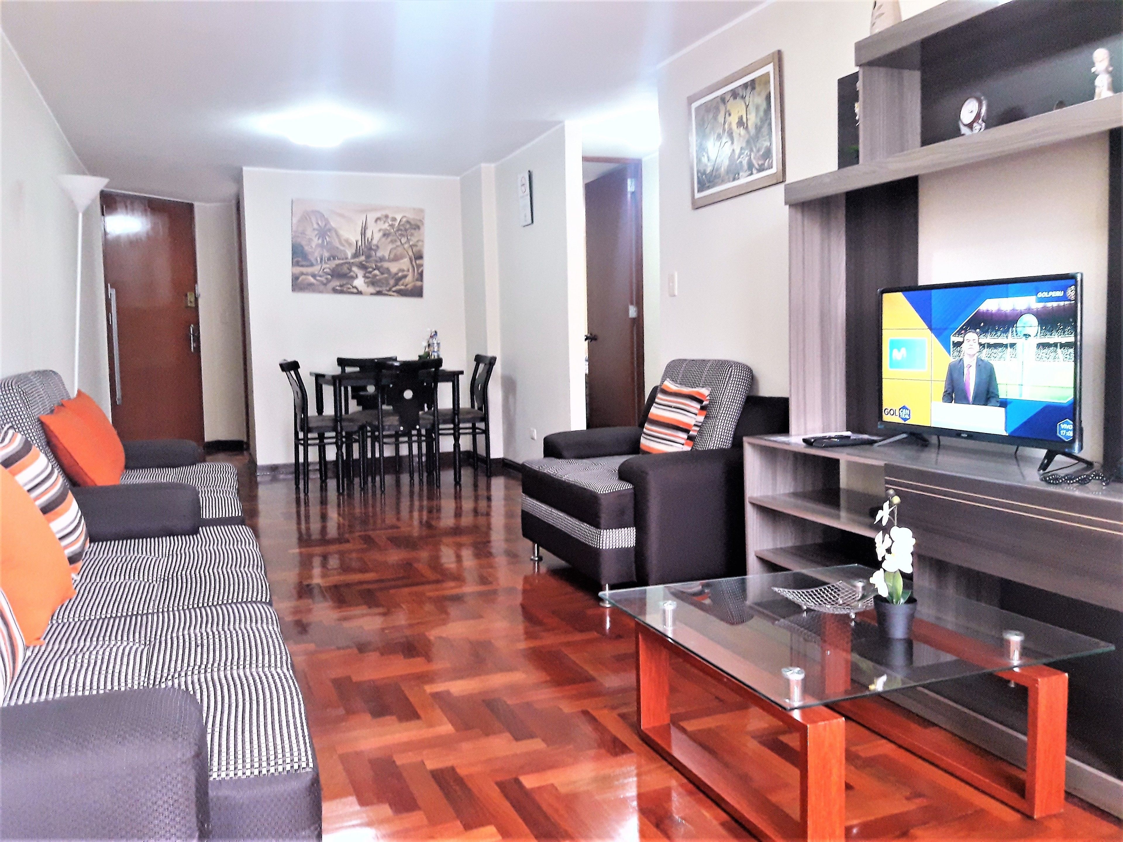 A living room with a sofa, a coffee table, a TV, and a dining area with chairs and a table.