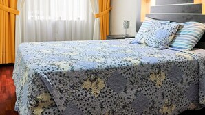 A neatly made bed with a floral bedspread, a bedside table with a lamp, and a window with curtains. - Eral Apartments Lima (Lima)