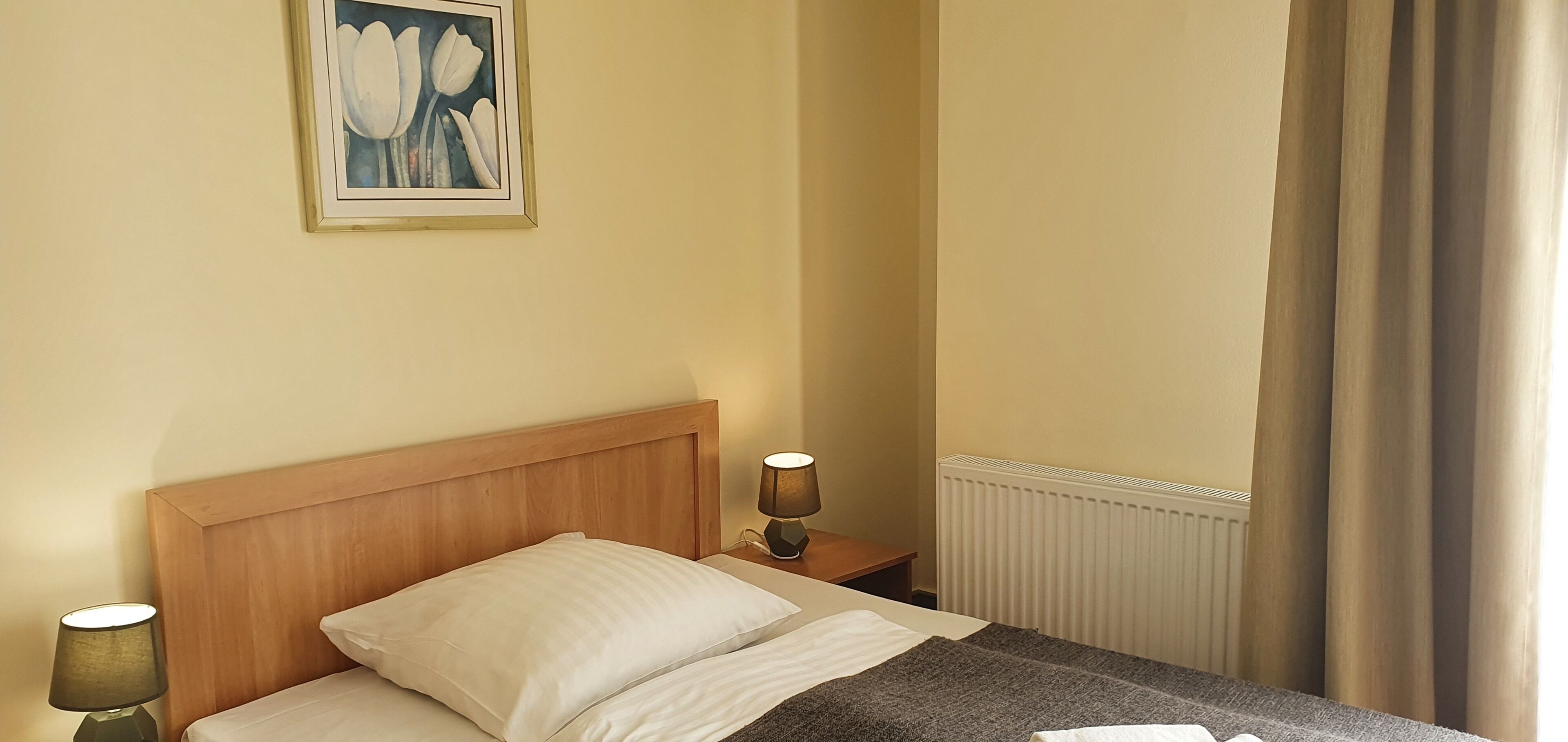 Een hotelkamer met een houten hoofdbord, een enkel bed met wit beddengoed, een bedlamp, een ingelijste afbeelding aan de muur, een radiator en een raam met gordijnen.