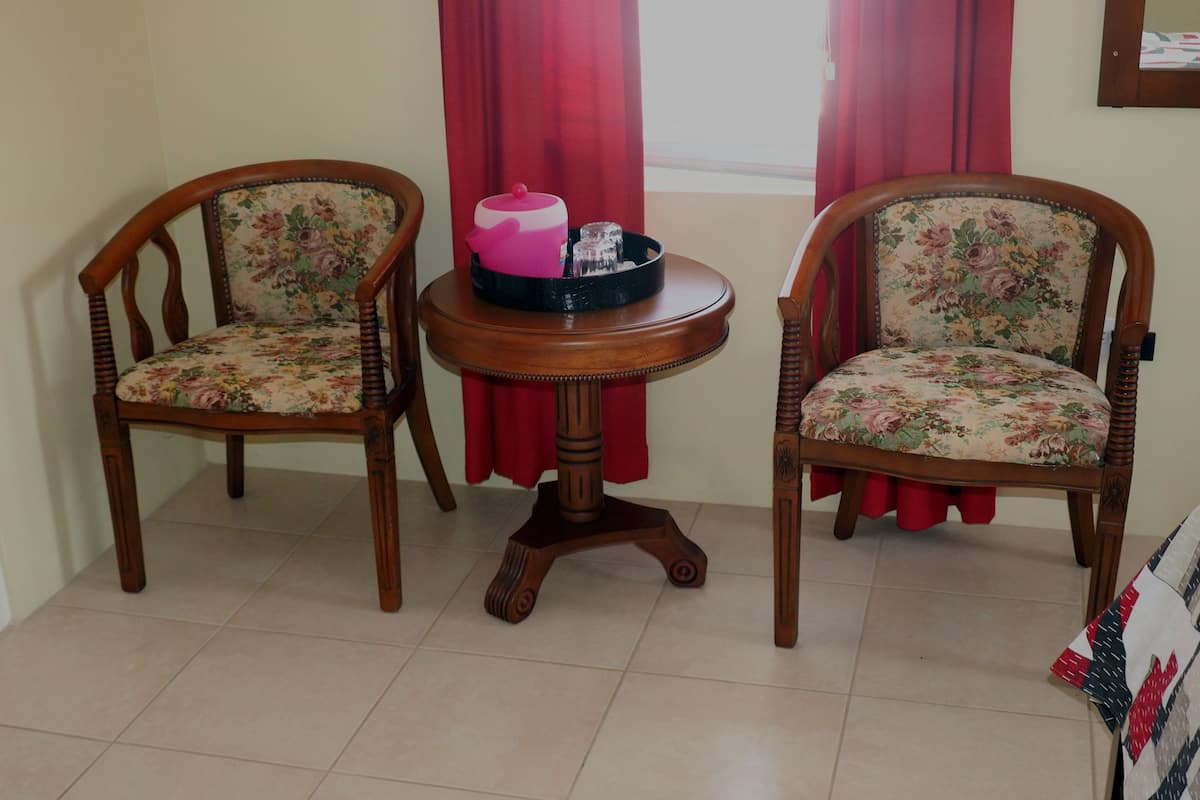 Dos sillas de madera con tapicería floral, una pequeña mesa de madera con un objeto rosa y un vaso, y una cortina roja al fondo.