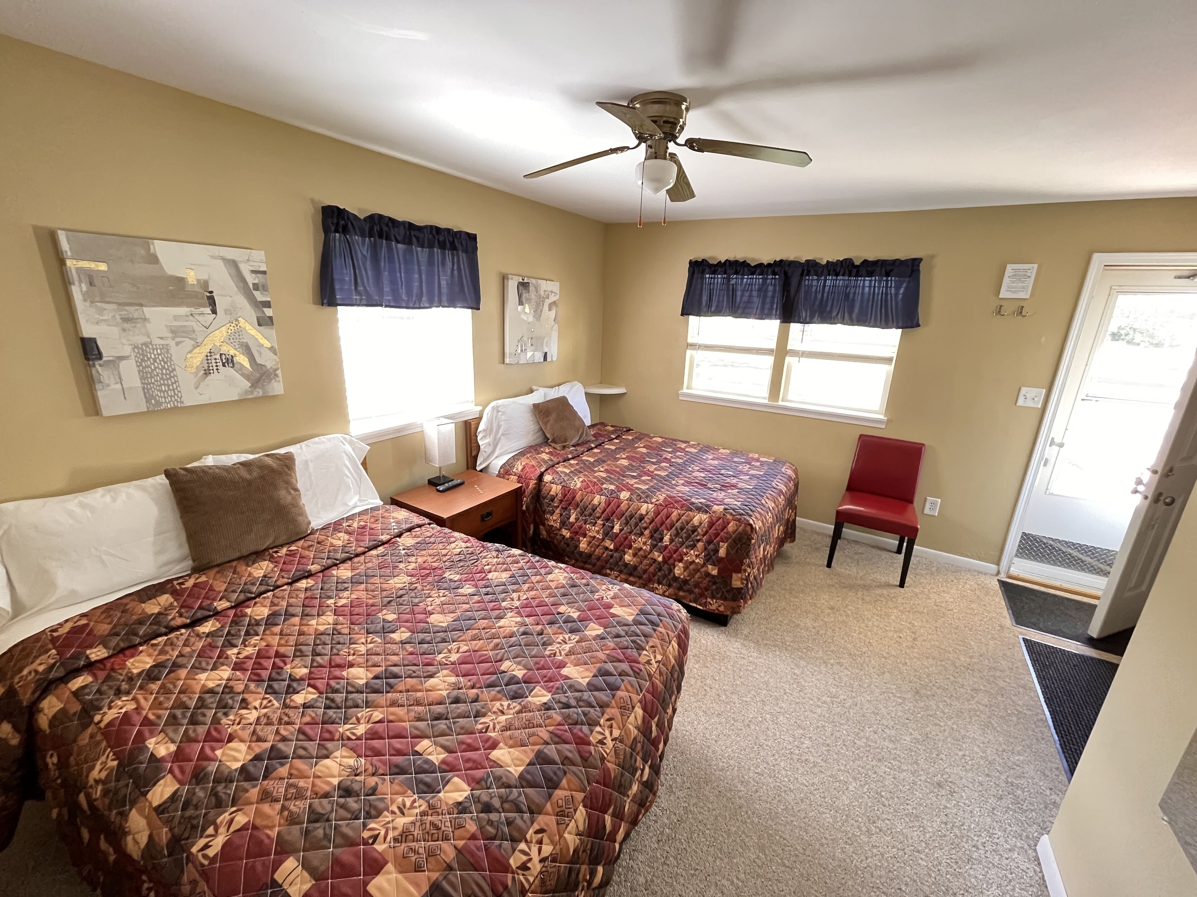 A hotel room with two beds, a ceiling fan, artwork on the walls, and a red chair.
