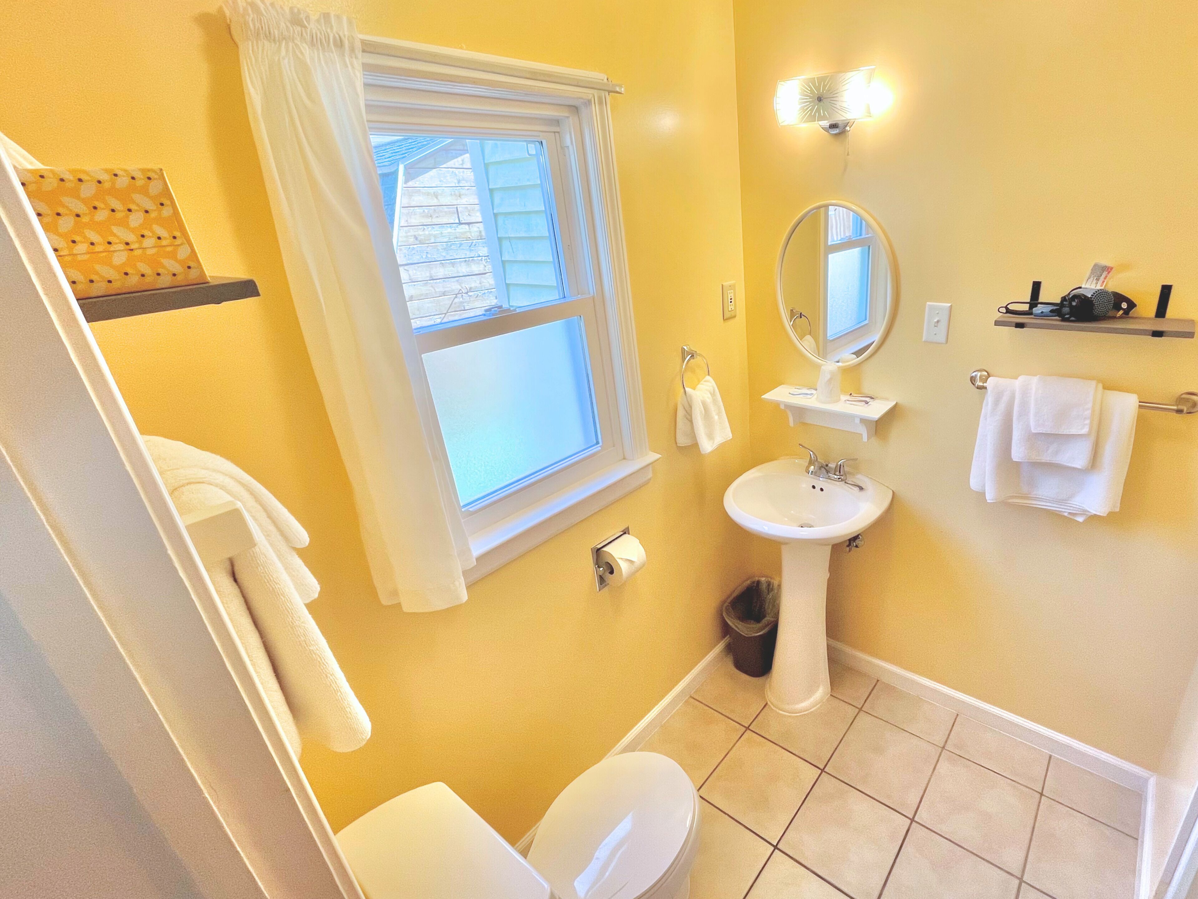 A bathroom with a toilet, sink, and window. There is a towel rack and a trash can.