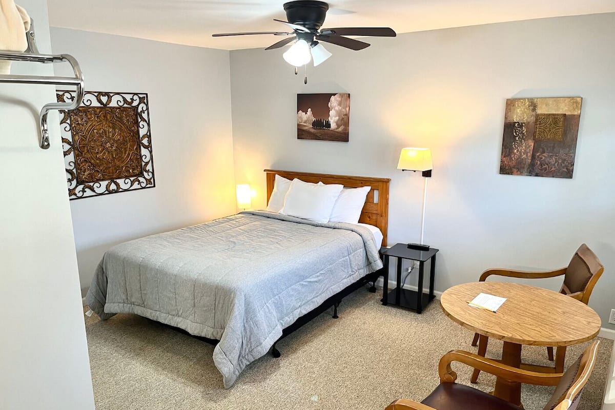 A hotel room with a bed, a round wooden table, two chairs, a ceiling fan, a wall-mounted lamp, and a framed picture.