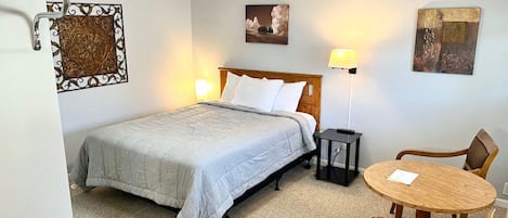 Et hotelværelse med en seng, et rundt træbord, to stole, en loftsvifter og en vægmonteret lampe samt et indrammet billede.