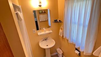 Een badkamer met een wastafel op een voetstuk, toilet en een spiegel boven de wastafel. Er is een handdoekhouder aan de muur en een prullenbak naast het toilet.