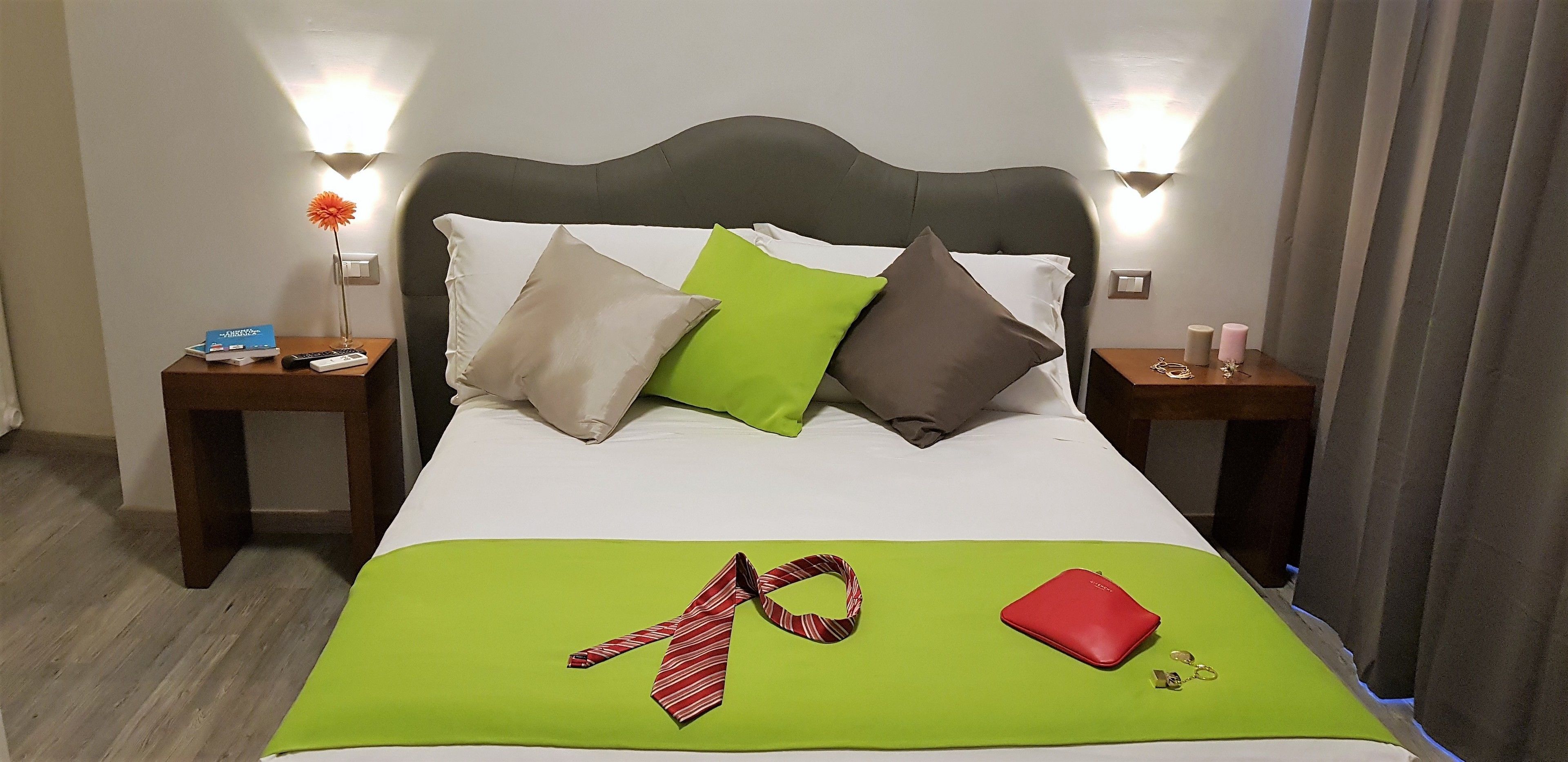 A hotel room with a neatly made bed, green and white bedding, a red tie, and a red wallet on the bed. There are two nightstands with lamps and a vase of flowers.