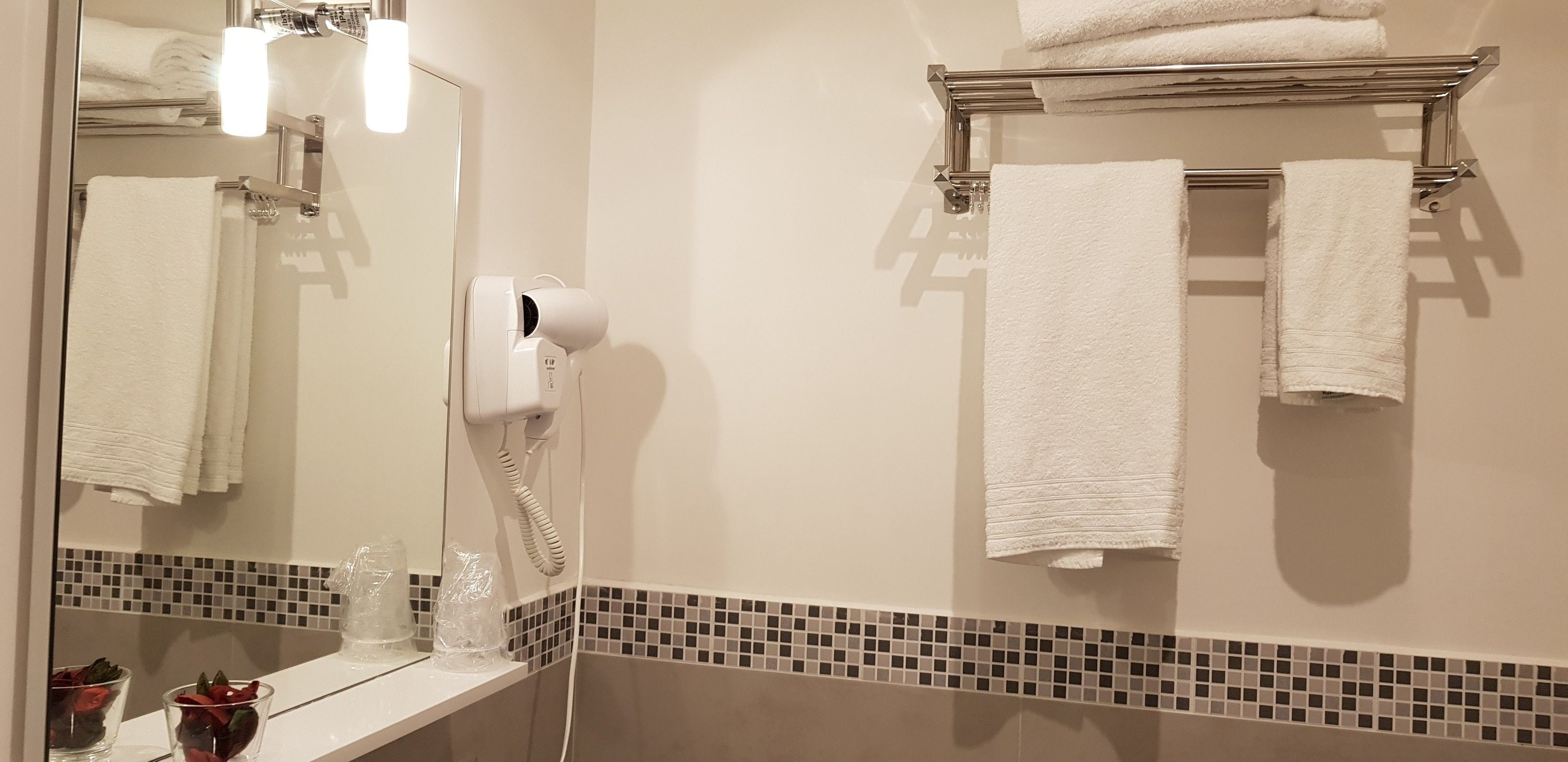 Un baño con espejo, toallero con toallas, secador de pelo y estante con vasos y una bolsa de plástico.