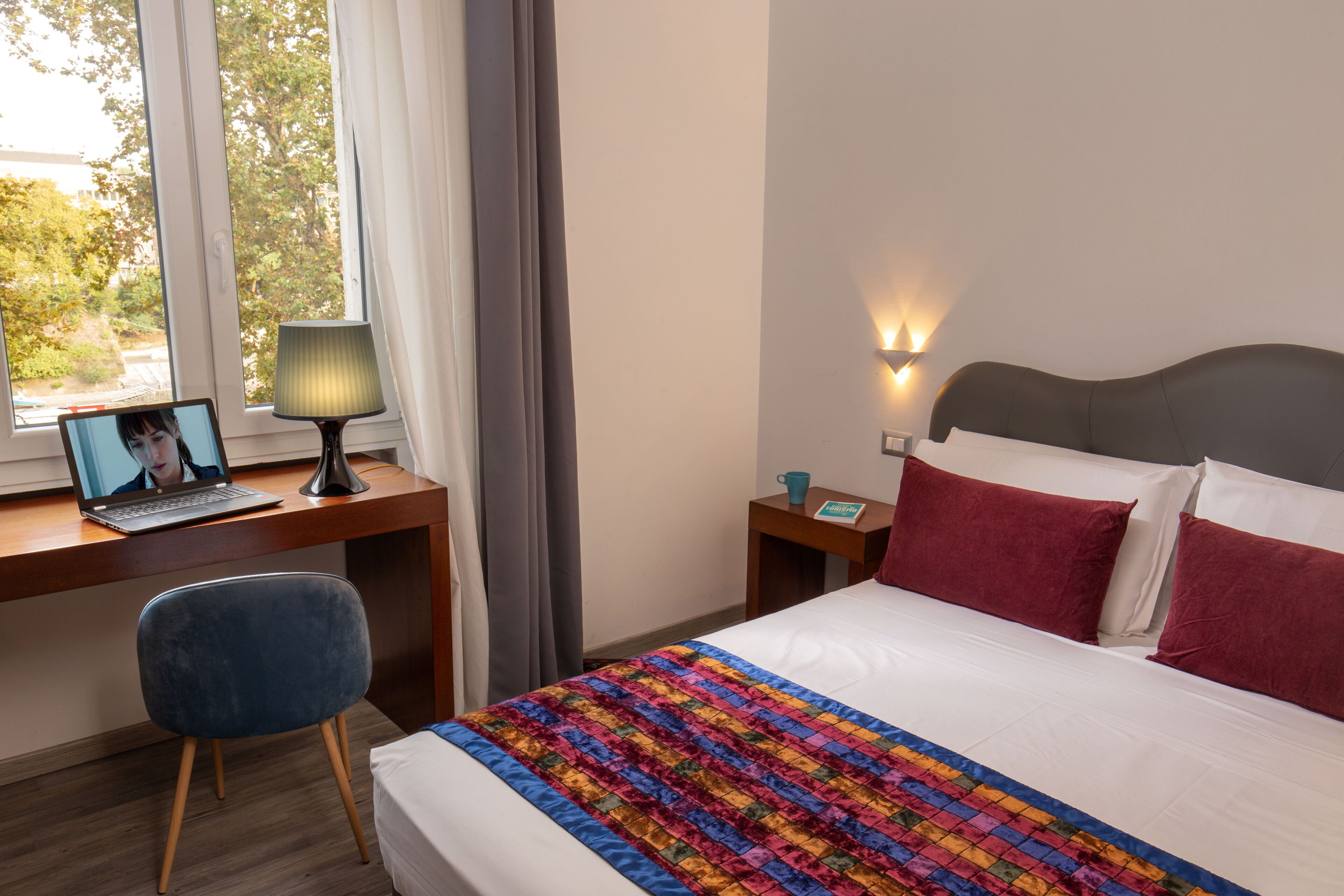 A hotel room with a bed, a desk with a laptop, a chair, a bedside table with a lamp, and a view of trees through the window.