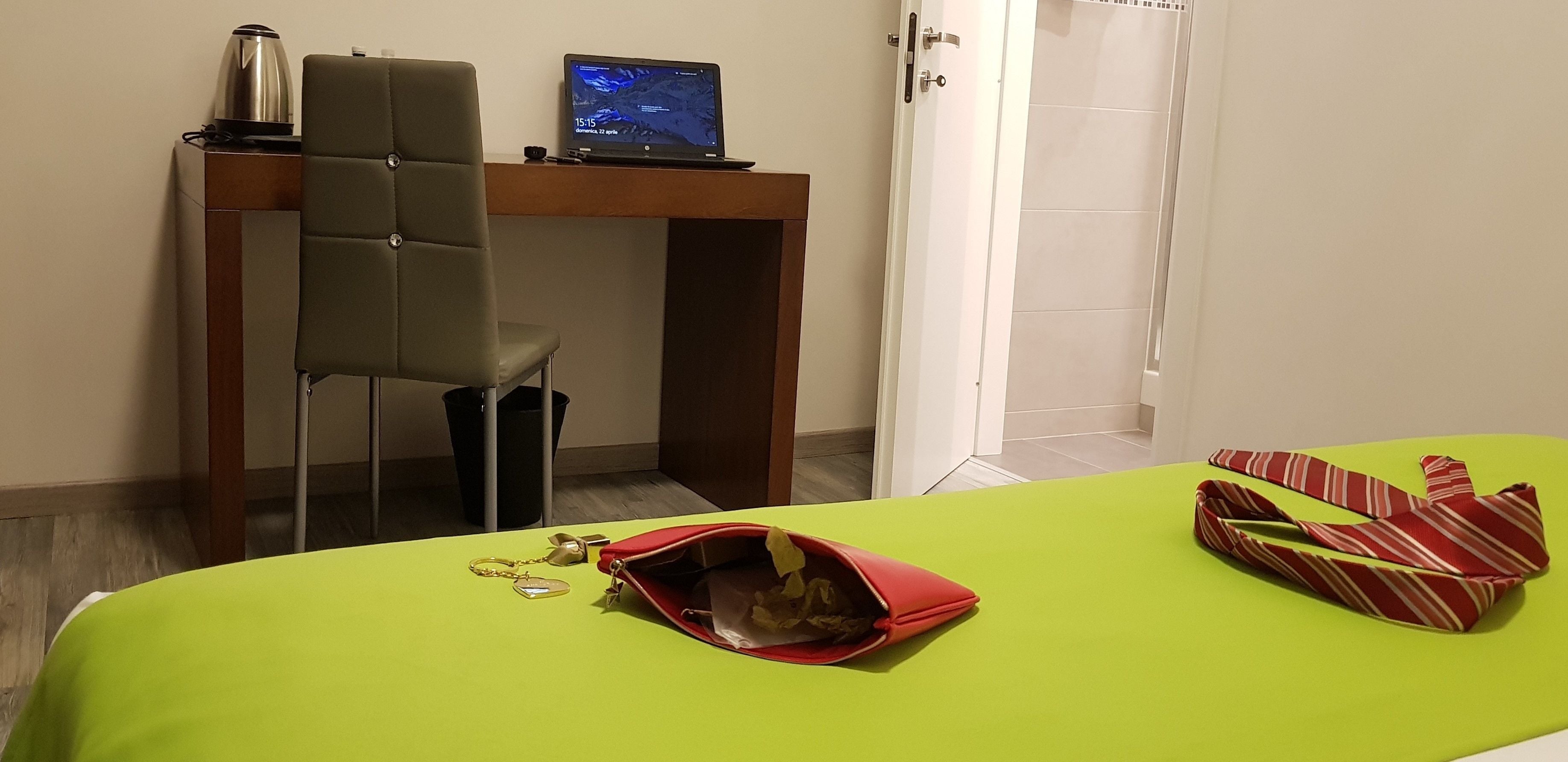 A hotel room with a desk, a laptop, a chair, a bed with a green bedspread, a red bag, and a striped tie.