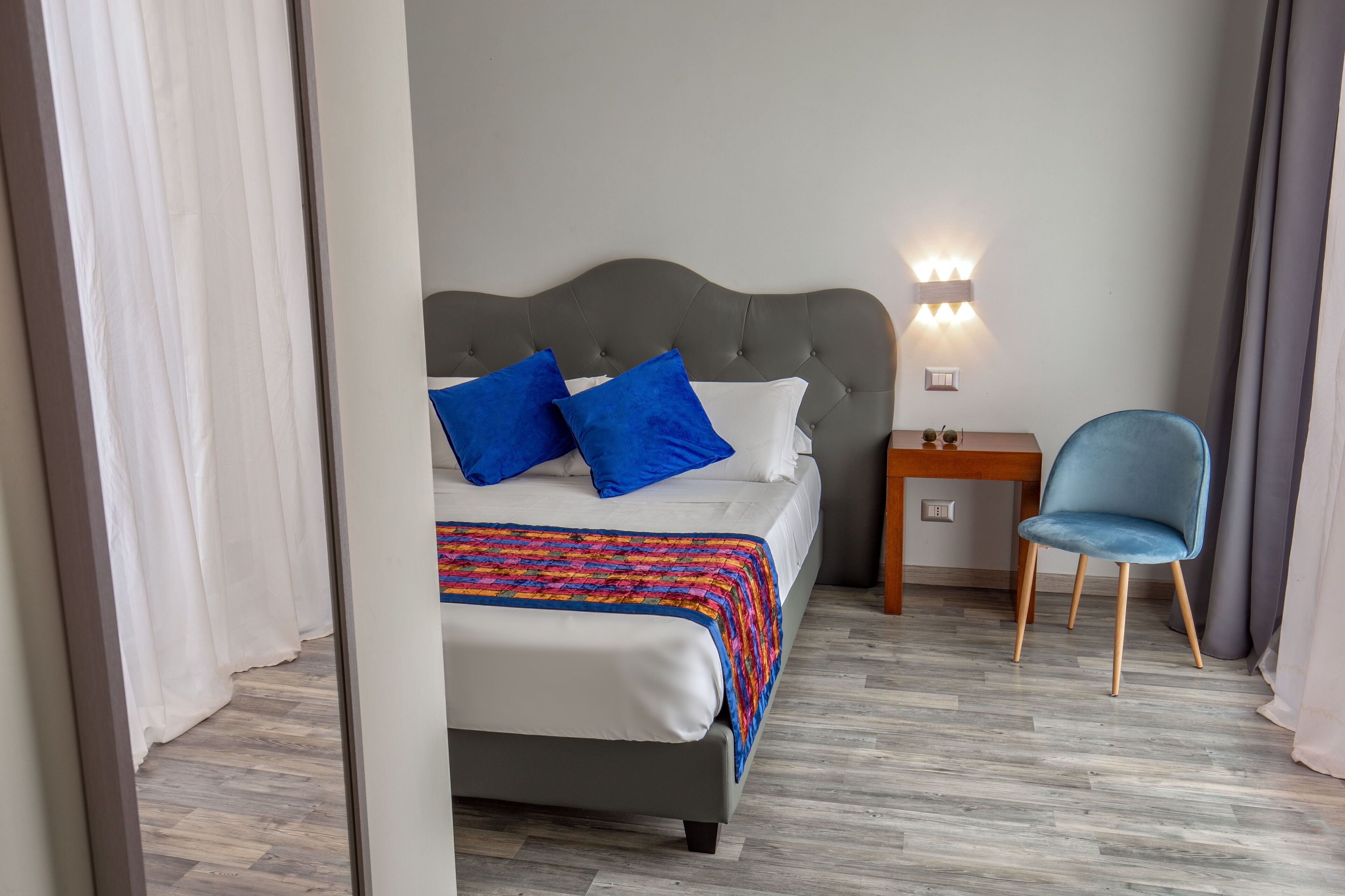 A neatly made bed with a grey headboard, a bedside table with a lamp, and a blue chair.