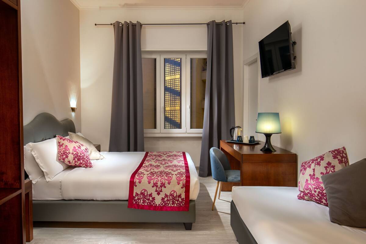 A hotel room with two beds, a desk, a TV, and a window with curtains.