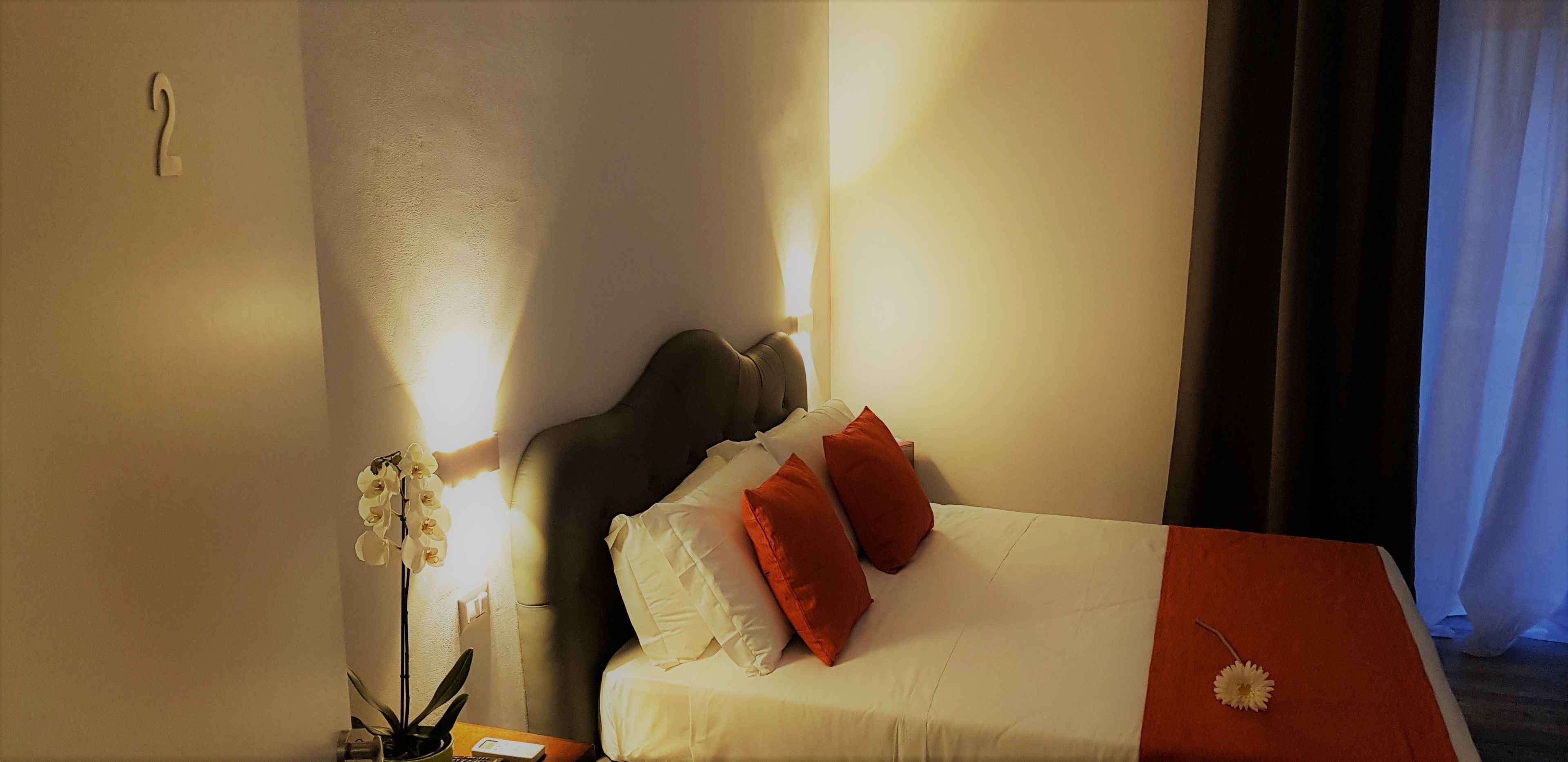 Ein Hotelzimmer mit Bett, Kissen, einem Nachttisch, einer Lampe und einer Orchideenpflanze.