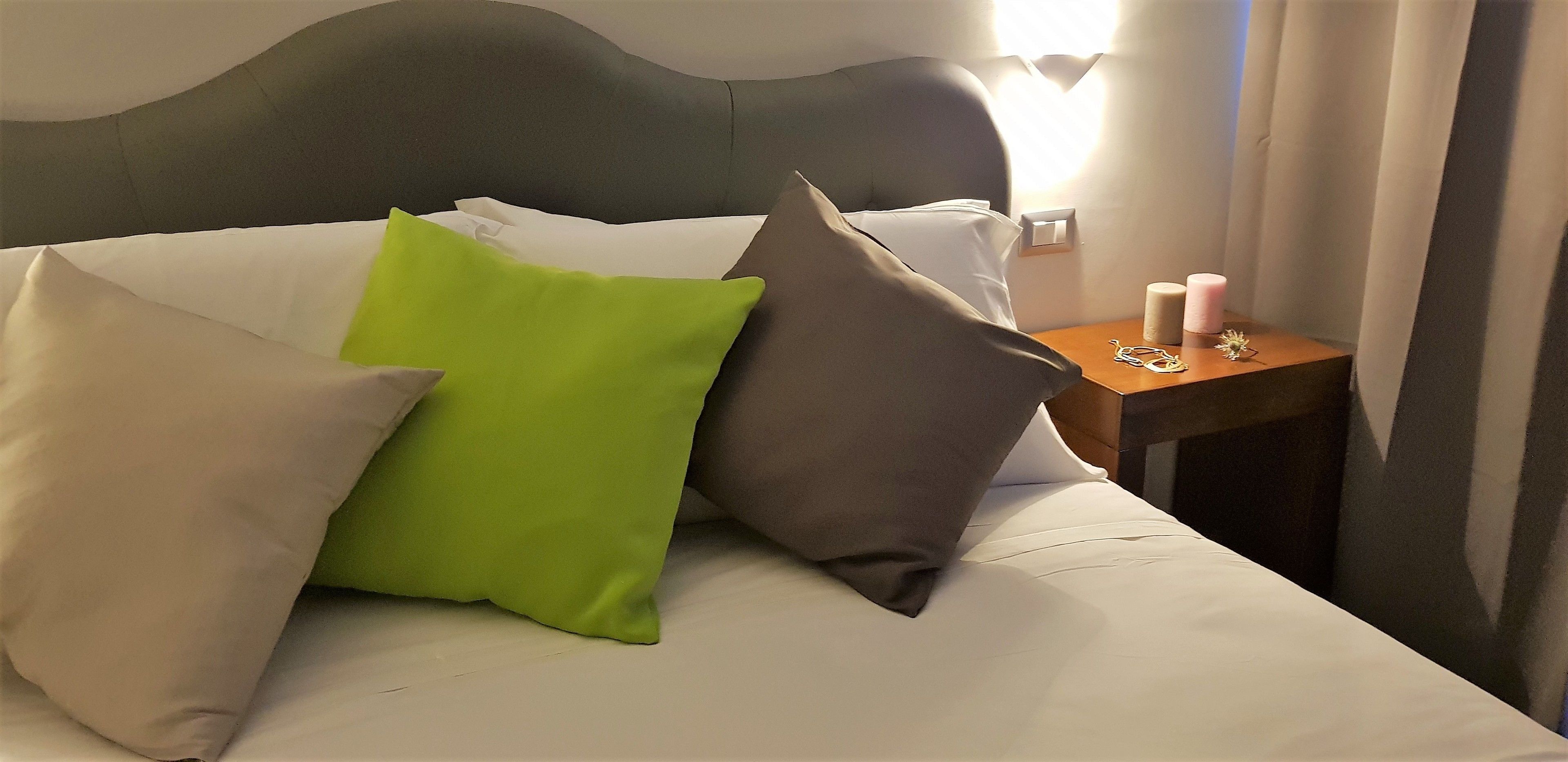 A neatly made bed with white and gray bedding, a green pillow, and a gray pillow. There is a wooden nightstand with a lamp and a small decorative item.