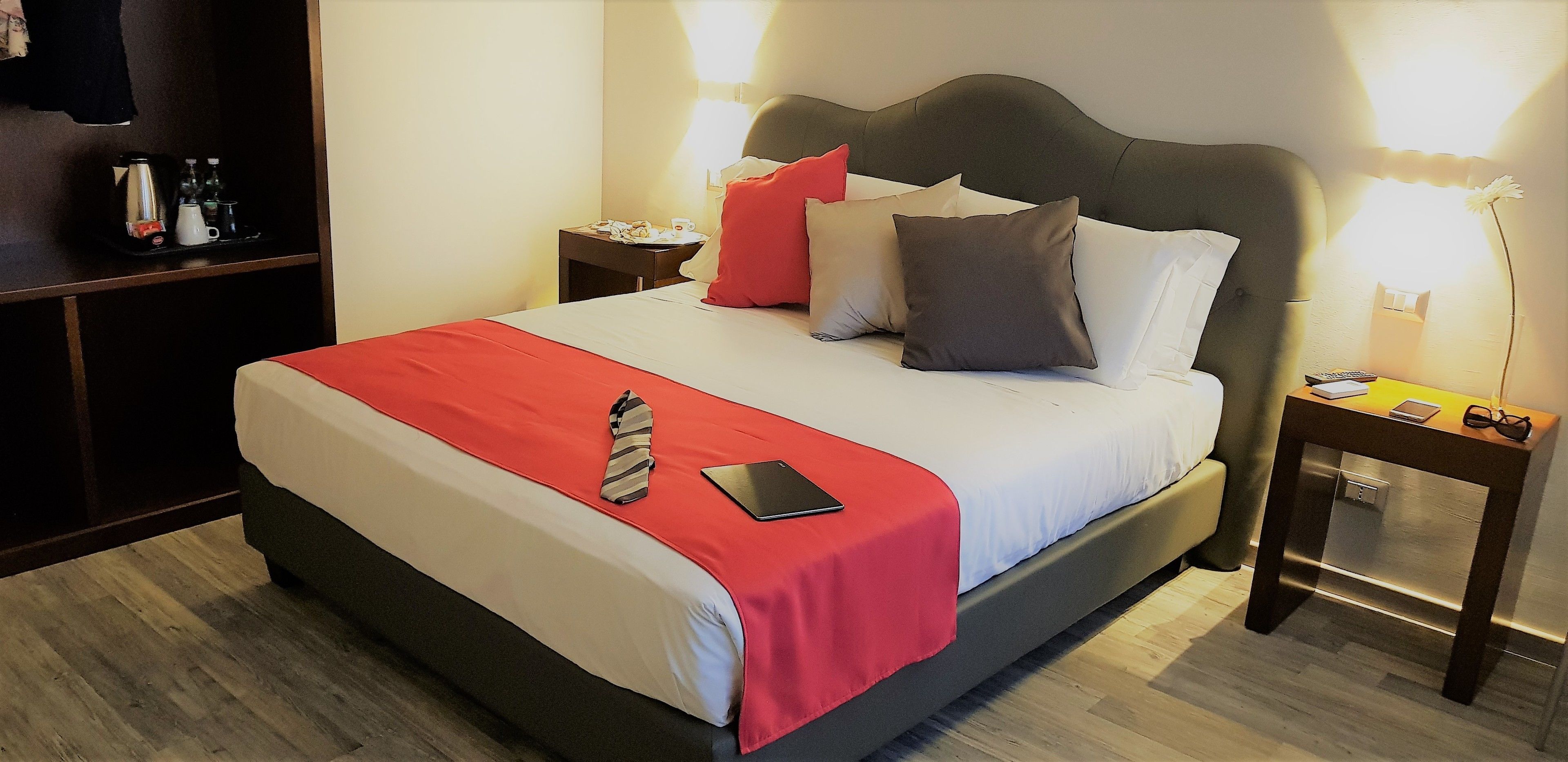 Una cama bien hecha con un edredón rojo, un teléfono inteligente y una corbata sobre la cama. Hay una mesita de noche con una lámpara y una bandeja con una cafetera y botellas.