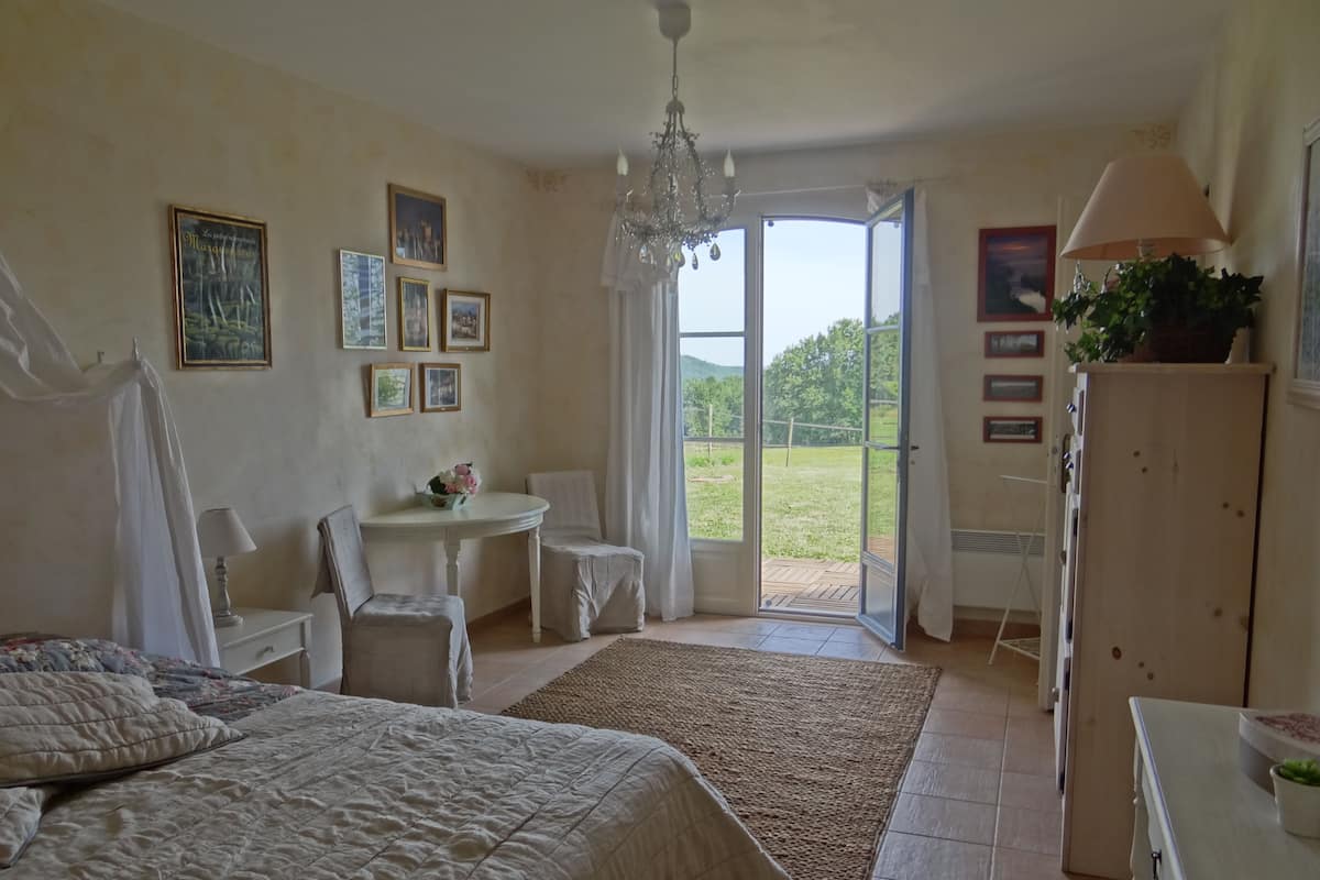 A bedroom with a bed, a desk, a chair, a chandelier, and a view of the outdoors.