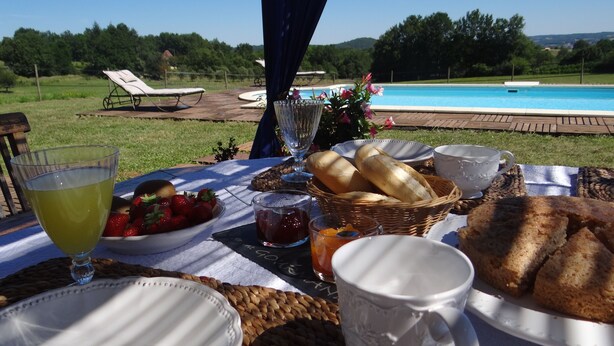 Mesa al aire libre con vistas a una piscina, con platos, pan, fresas y un vaso de jugo.