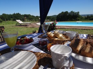 A table set outdoors with a view of a swimming pool, featuring plates, bread, strawberries, and a glass of juice. - La Rolandie haute (Limeuil)