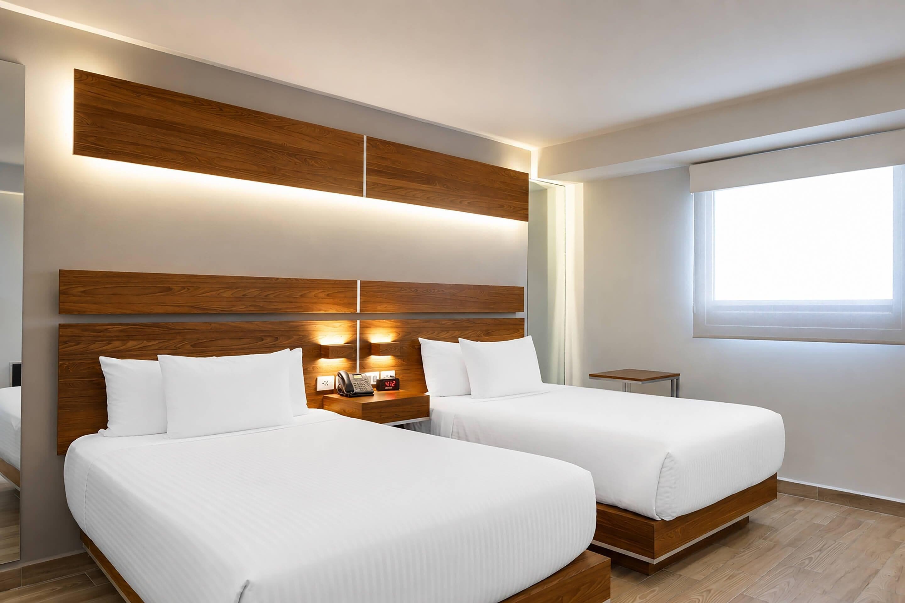 A hotel room with two beds, a bedside table with a phone and clock, and a window with curtains.