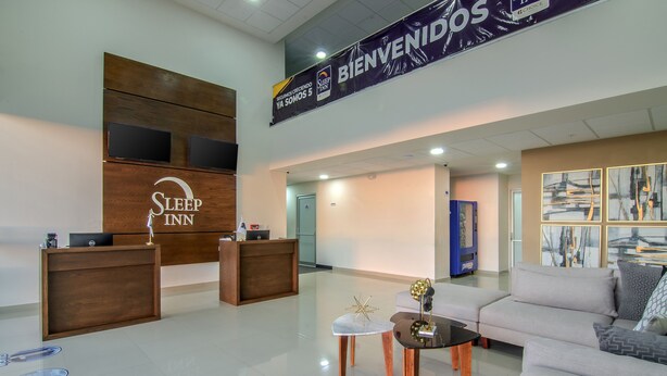 Un moderno vestíbulo de hotel con una recepción, una zona de estar con sofá y mesa de centro, y obras de arte en las paredes.