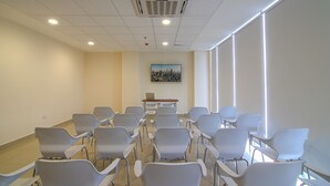 Un salón de conferencias con hileras de sillas, una mesa con una computadora portátil y un cuadro de un paisaje urbano en la pared.