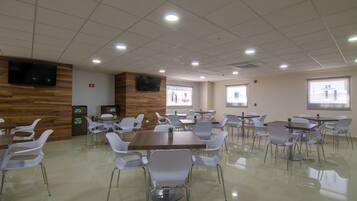 A spacious cafeteria with multiple tables and chairs, a wooden accent wall, a mounted TV, and windows providing natural light.