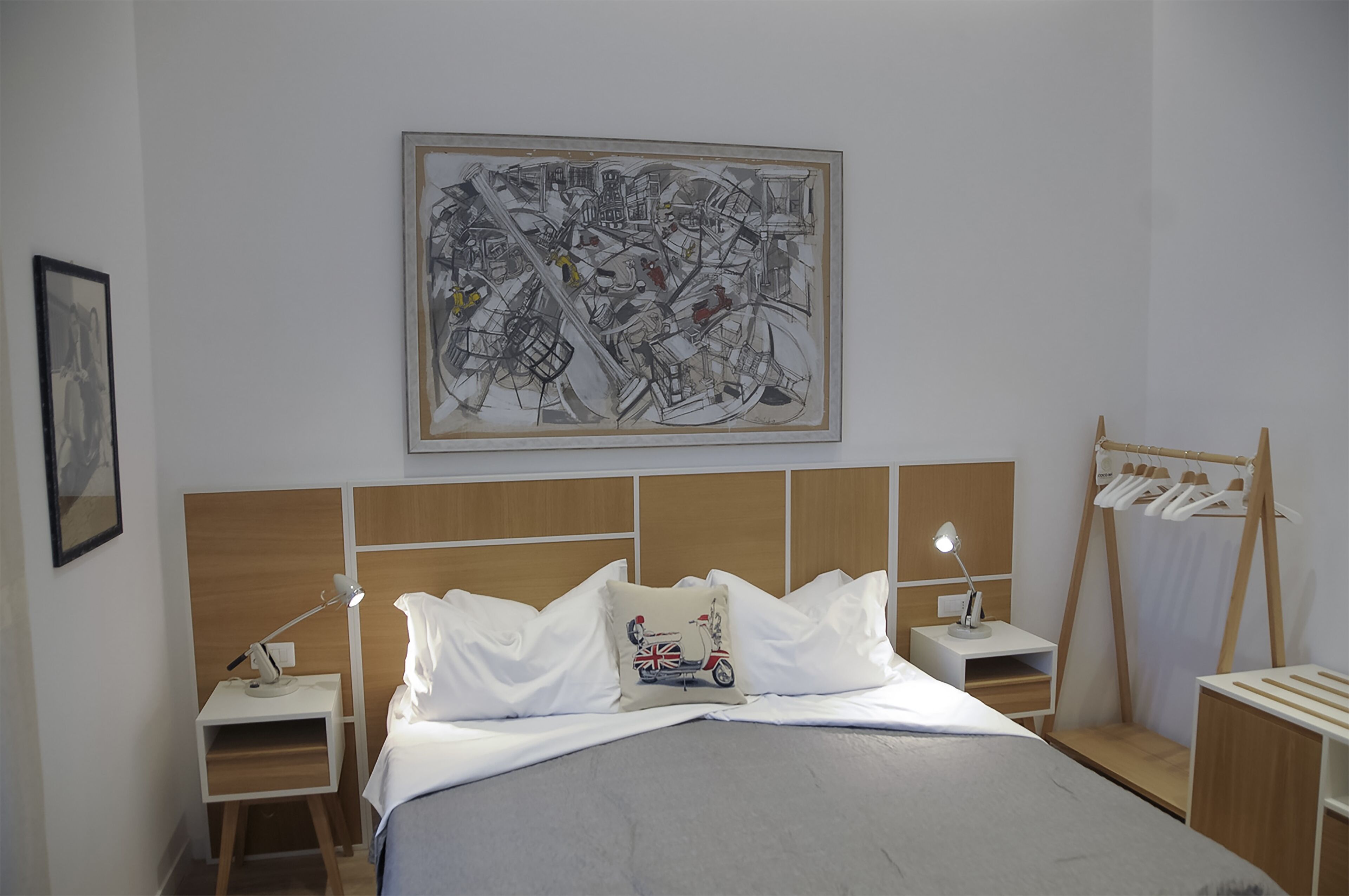 A neatly arranged hotel room with a large bed, a bedside table with a lamp, a wall-mounted artwork, and a clothing rack.