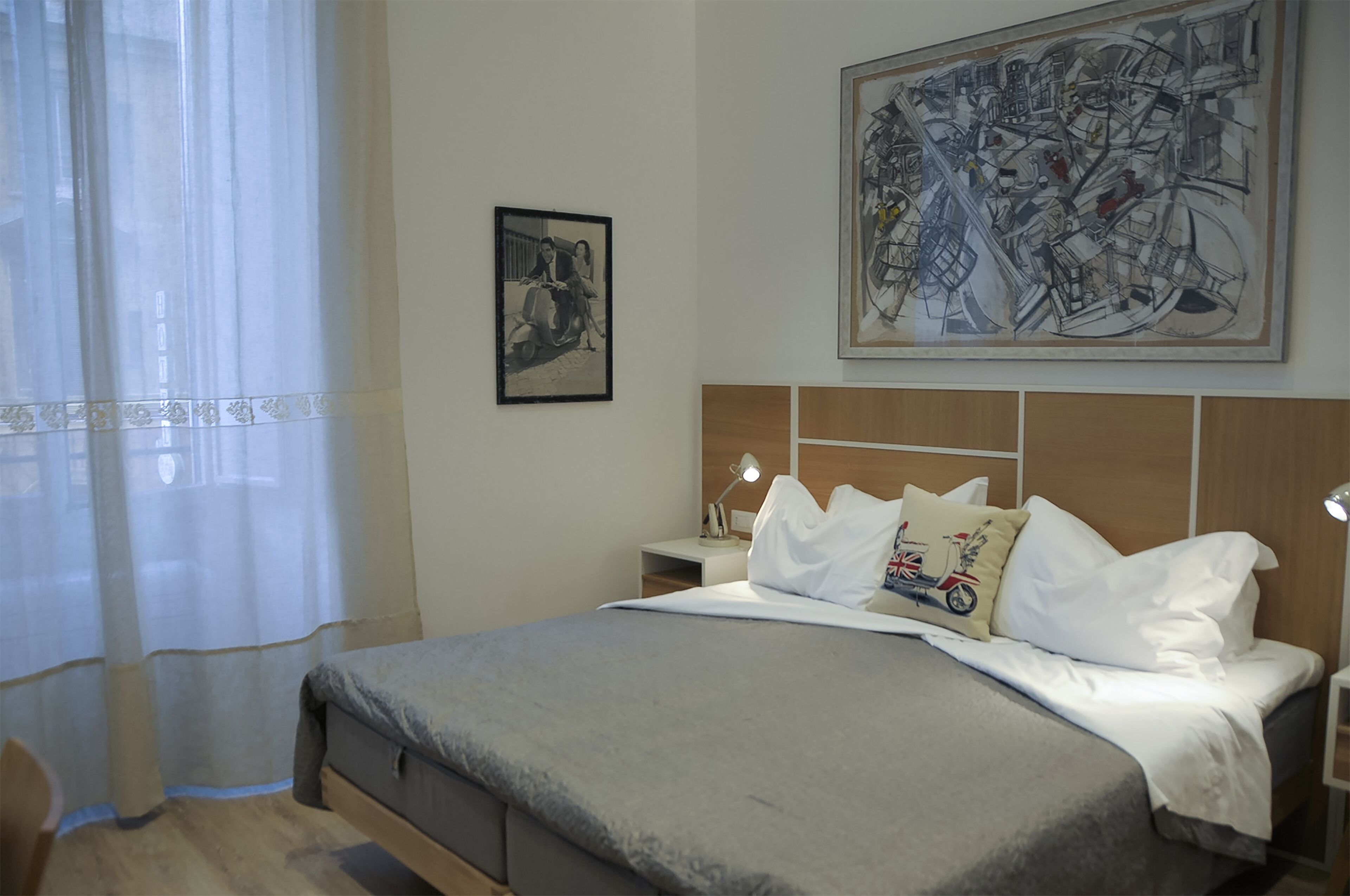A neatly made bed with a grey bedspread, a pillow with a motorcycle design, and a headboard with a wooden panel. There is a framed picture on the wall and a lamp on the bedside table.