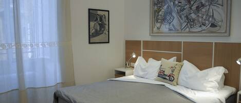 A neatly made bed with a grey bedspread, a pillow with a motorcycle design, and a headboard with a wooden panel. There is a framed picture on the wall and a lamp on the bedside table.