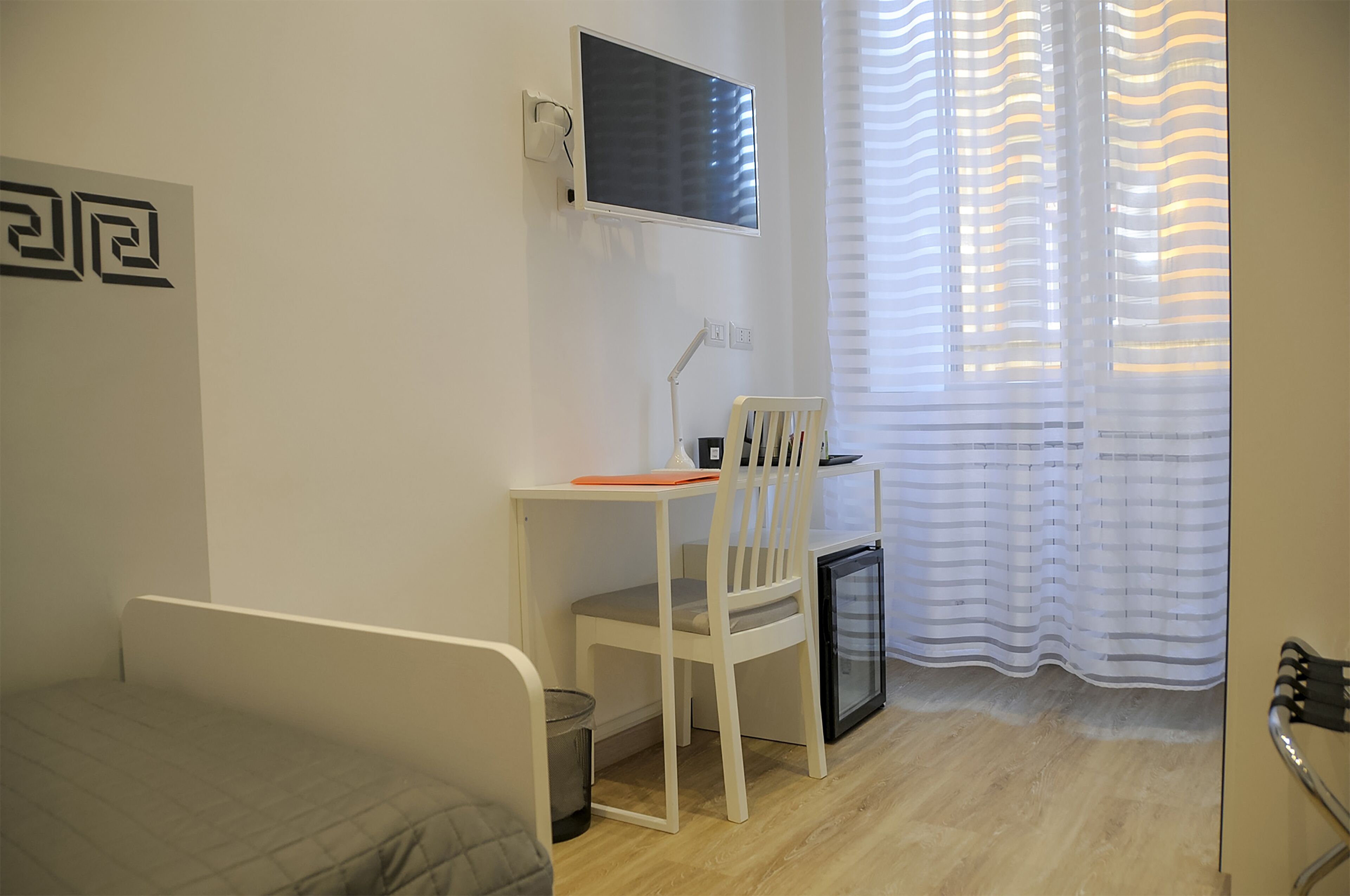 Une petite pièce propre et bien rangée, avec un lit, un bureau et une chaise, une télévision et un petit réfrigérateur.