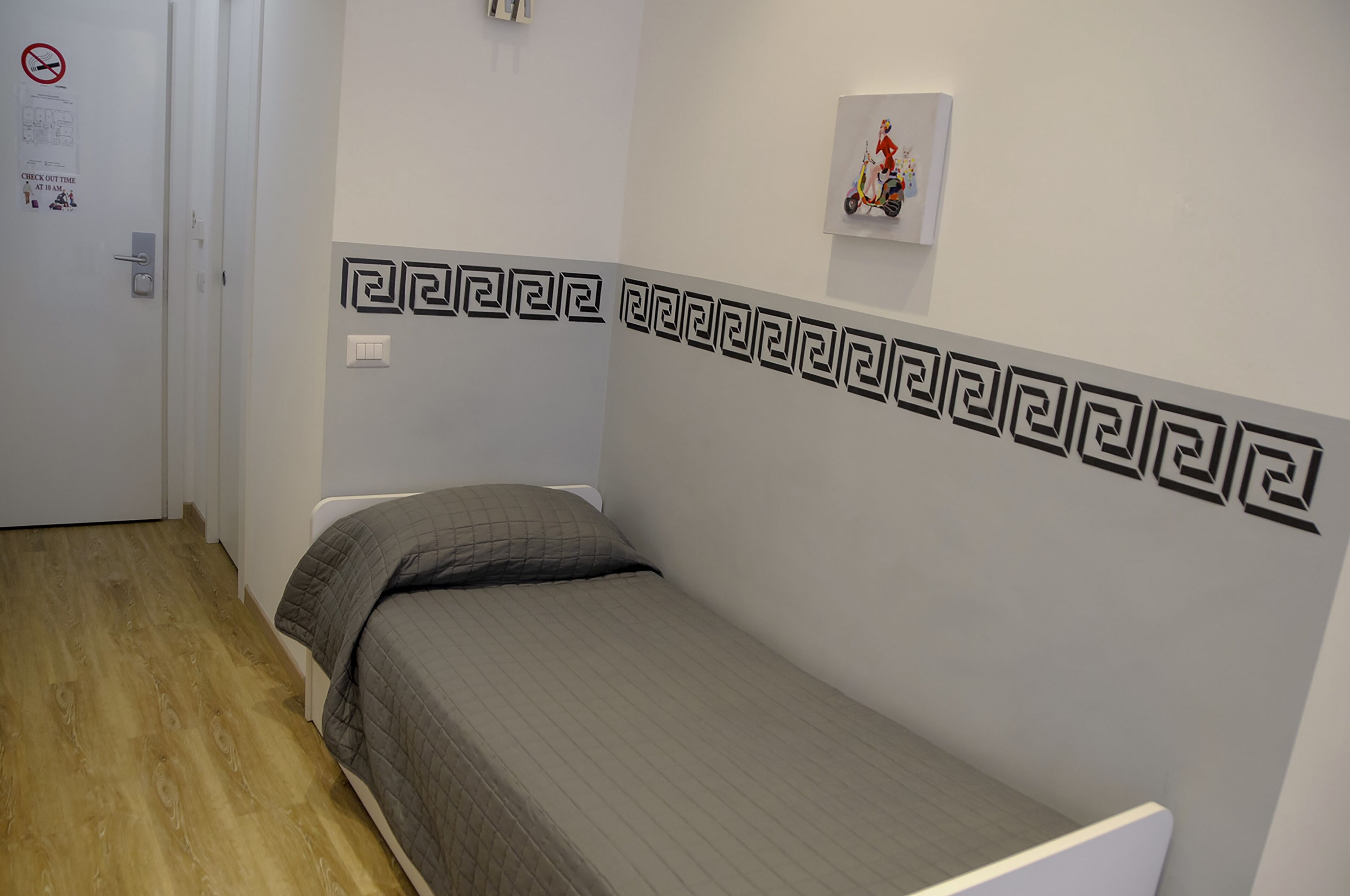 A small, single-bed room with a wooden floor, a grey bedspread, and a decorative wall pattern.