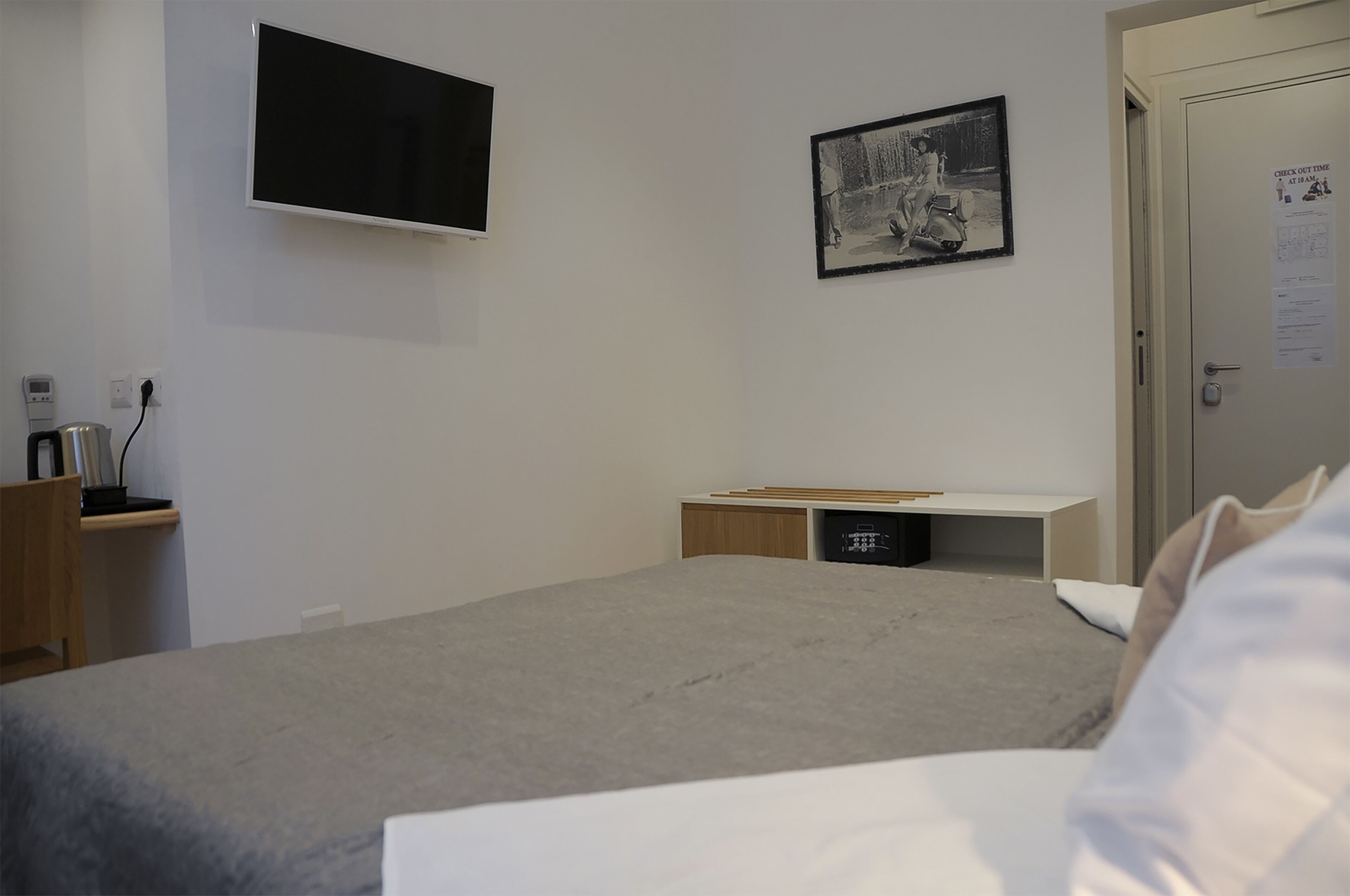 A bedroom with a bed, a desk with a chair, a TV mounted on the wall, and a door with a notice.