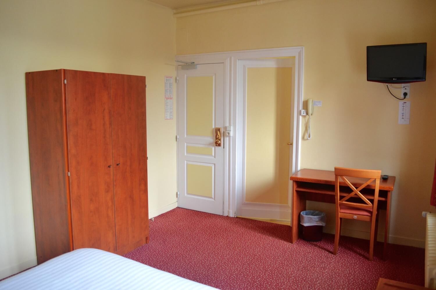 Een kamer met een bed, een houten garderobe, een bureau met stoel, een prullenbak en een aan de muur bevestigde televisie.